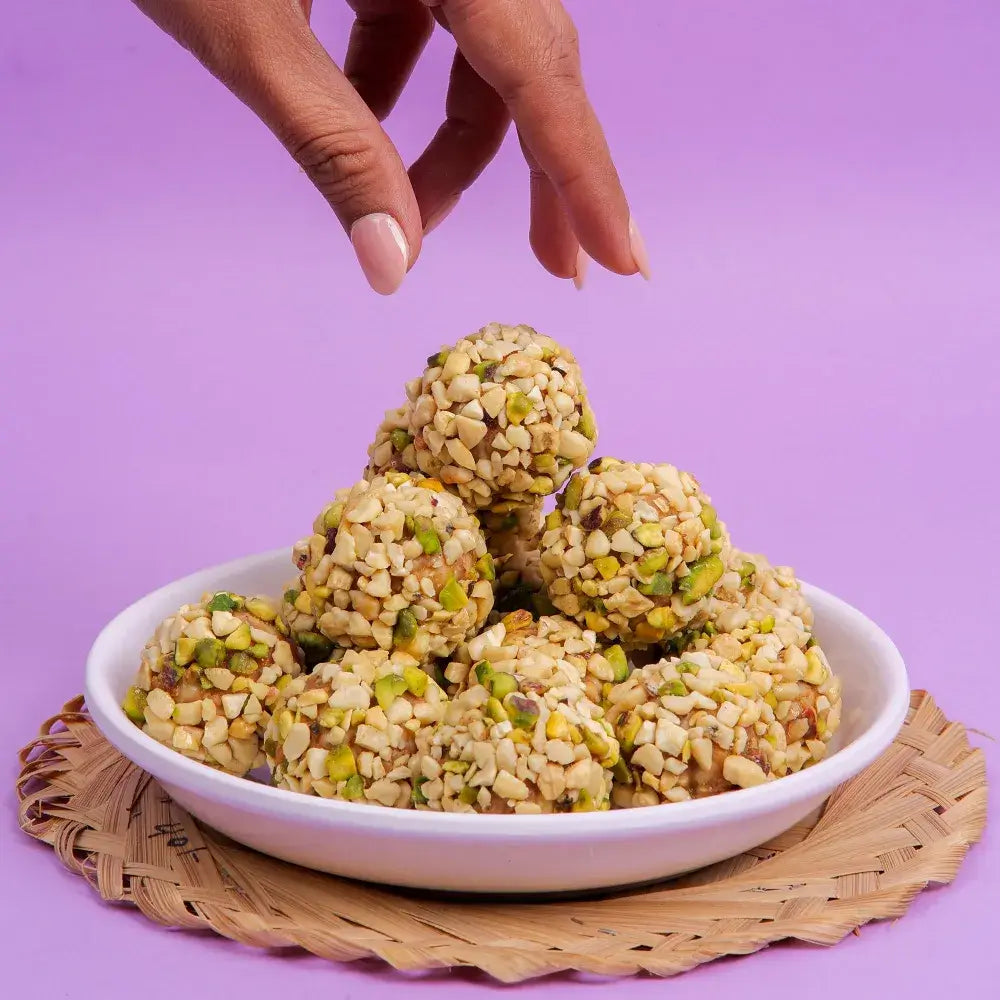 Best kaju katli and ladoo in Surat, featuring a hand picking a Kaju Pista Ladoo from a white plate on a purple background.